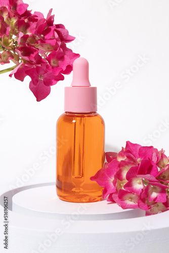 Cosmetic glass bottle with dropper for essential oils and serum on a podium with pink flowers. Face and body care spa concept and natural cosmetics