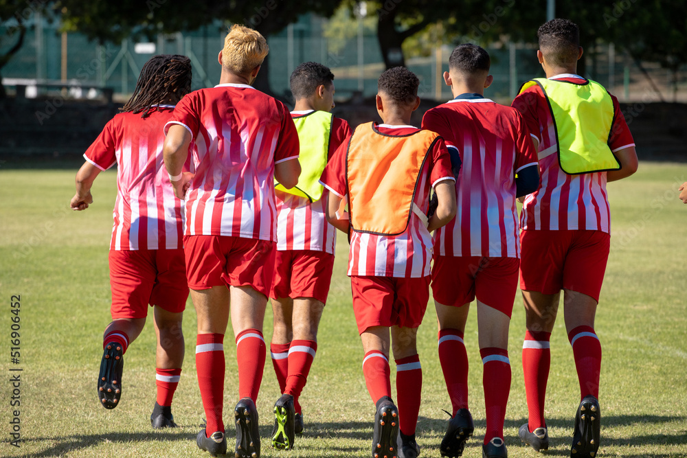 Rear view of multiracial male soccer players wearing red sports uniforms running at playground Rear view of multiracial male soccer players wearing red sports uniforms running at playground