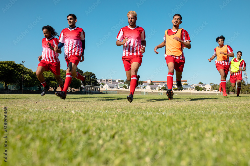 Obraz premium Full length of multiracial male athletes in red uniforms running on grassy land against clear sky