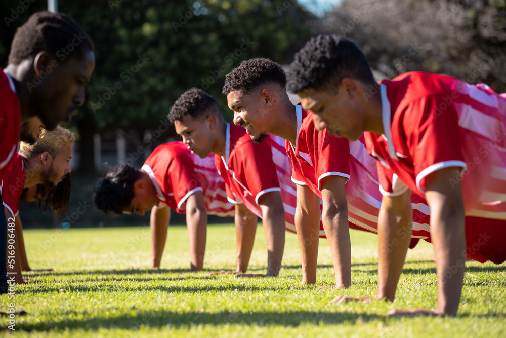 Fototapeta premium Male multiracial players in red uniforms practicing push-ups in a row on grassy field at playground