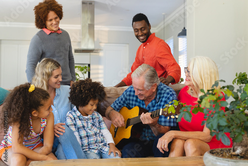 Multiracial happy multigeneration family looking at senior man playing guitar in living room