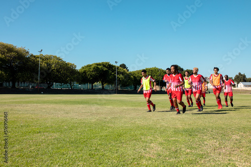 Multiracial male soccer team wearing red uniforms running on grassy land against clear sky