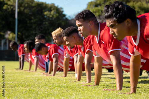 Male multiracial players wearing red uniforms in a row doing push-ups on grassy playground