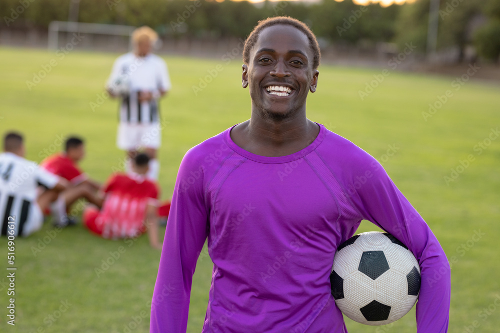 Portrait of happy african american goalkeeper with soccer ball standing in playground at sunset