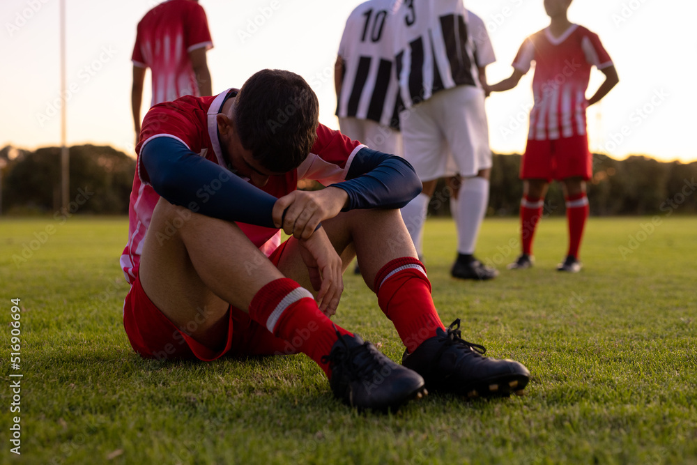 Caucasian sad male athlete sitting on grassy land with team players in background at playground Caucasian sad male athlete sitting on grassy land with team players in background at playground