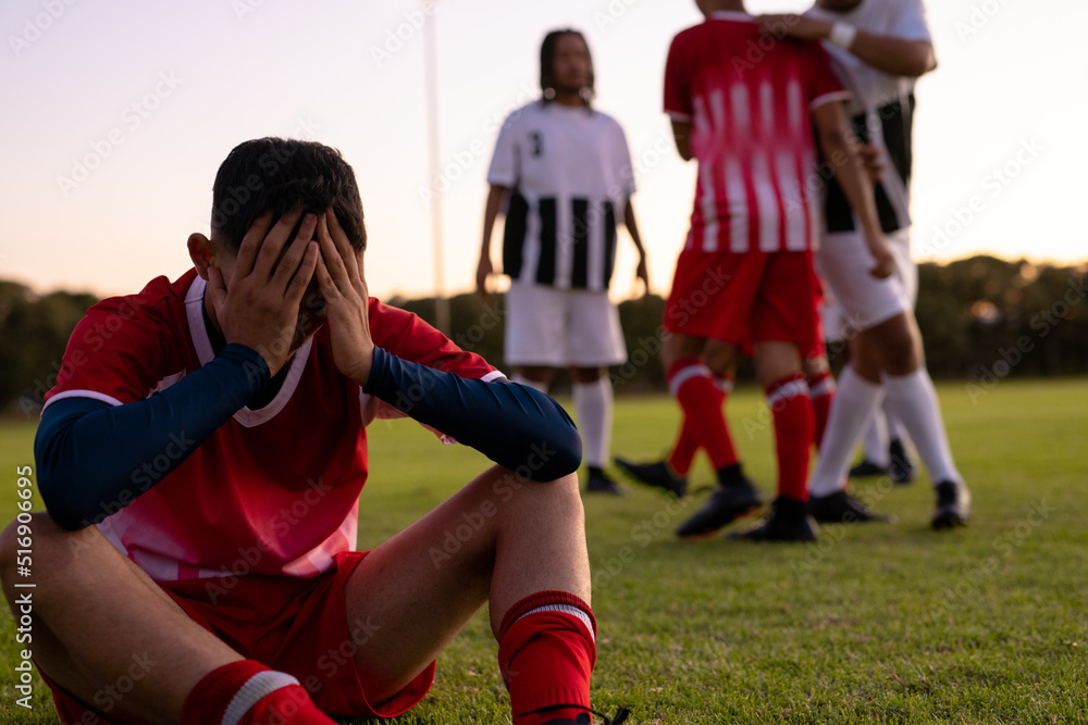 Multiracial sad male soccer player with head in hands sitting on grassy land against clear sky Multiracial sad male soccer player with head in hands sitting on grassy land against clear sky
