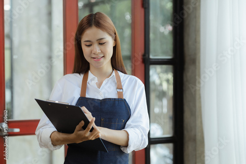Asian woman in an apron stands in front of the door holding the restaurant's menu to welcome customers, the opening of a small restaurant, a woman operating. Operating a small restaurant.