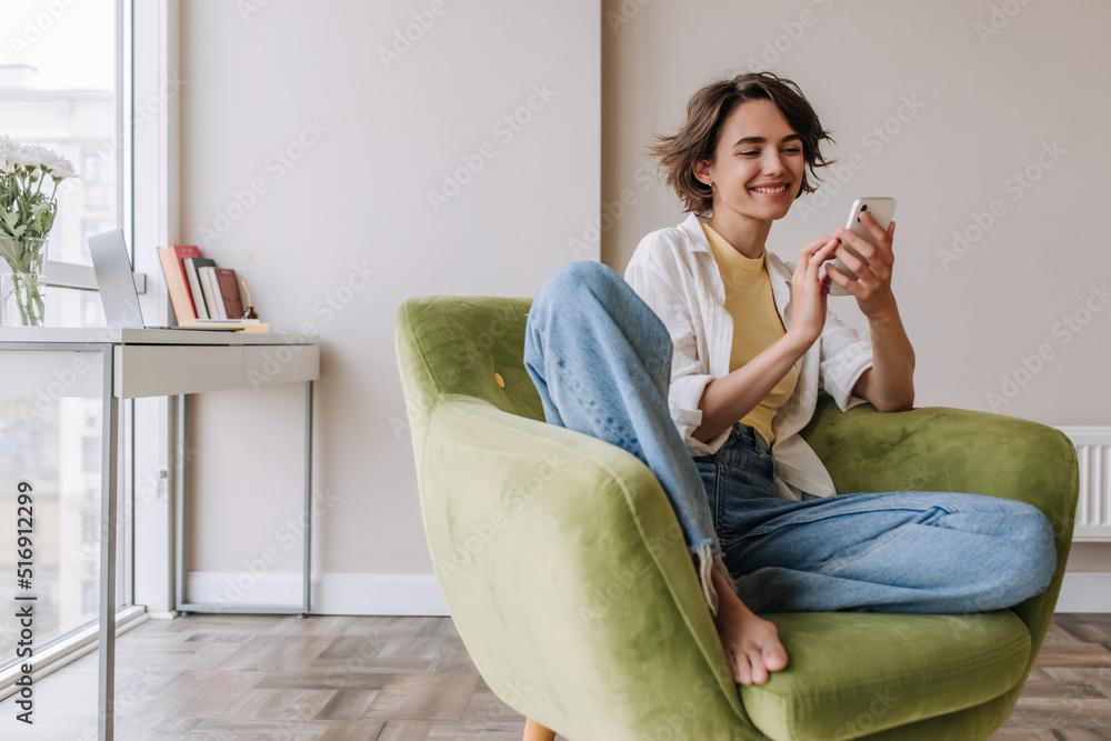 Caucasian brunette woman smiling at phone . Full view of pretty young woman holding and chatting on smartphone in home. Technology, lifestyle Caucasian brunette woman smiling at phone . Full view of pretty young woman holding and chatting on smartphone in home. Technology, lifestyle