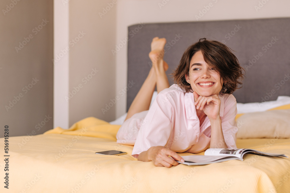 Brunette young woman on the bad smiling . Wearing pink pajamas lying and looking away with magazine and phone. Concept of leisure life, relaxing Brunette young woman on the bad smiling . Wearing pink pajamas lying and looking away with magazine and phone. Concept of leisure life, relaxing