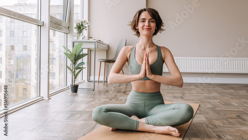 Cheerful young brunette woman sitting on lotus yoga pose . Caucasian fair skinned girl smiling and looking to camera in home dressed green top and leggings. Workout, leisure life 