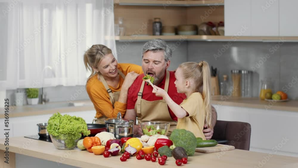 Cute little girl feeding father with freshly made vegetable salad, loving man enjoying meal, tracking shot, slow motion Cute little girl feeding father with freshly made vegetable salad, loving man enjoying meal, tracking shot, slow motion