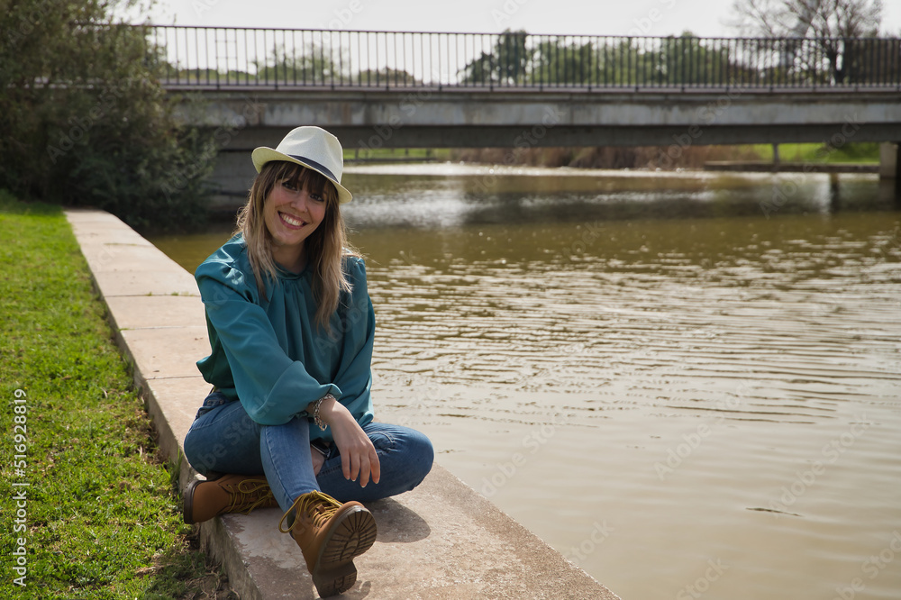 Young, beautiful, blonde woman in green shirt, jeans and hat, sitting smiling by a river. Concept warmth, sun, spring, summer, vacation.