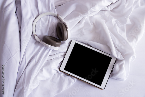 a tablet and headphones lie on a crumpled white bed, top view, close-up