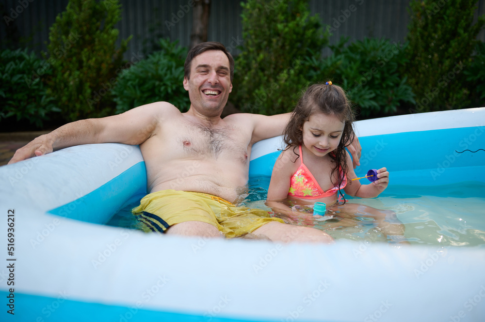Happy young father is having fun with his little daughter blowing soap balls in the inflatable pool on a warm summer day