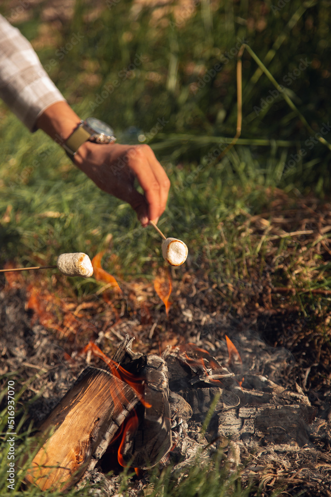 the guy's hand holds a white marshmallow on a stick over a fire, fry marshmallows in nature
