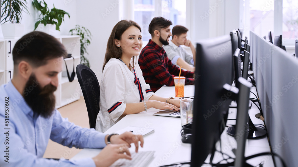 Junior female professional working in modern IT office. Group of young and experienced programmers and software developers sitting at desks working on computers.
