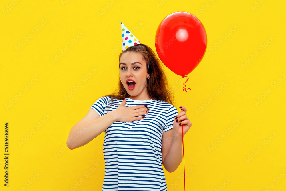 Surprised young woman in a birthday cap holds hand on chest and holds red inflated helium balloon, celebrates birthday or special occasion on a yellow background. Holiday and party concept