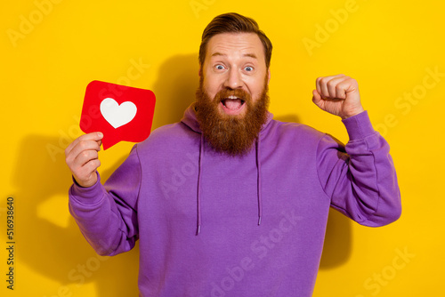 Photo of positive lucky guy dressed purple sweater holding love heart card rising fist isolated yellow color background © deagreez