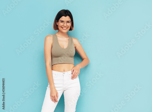 Young beautiful smiling female in trendy summer jeans clothes. Carefree woman posing near blue wall in studio. Sexy positive model having fun indoors. Cheerful and happy. Isolated. In glasses
