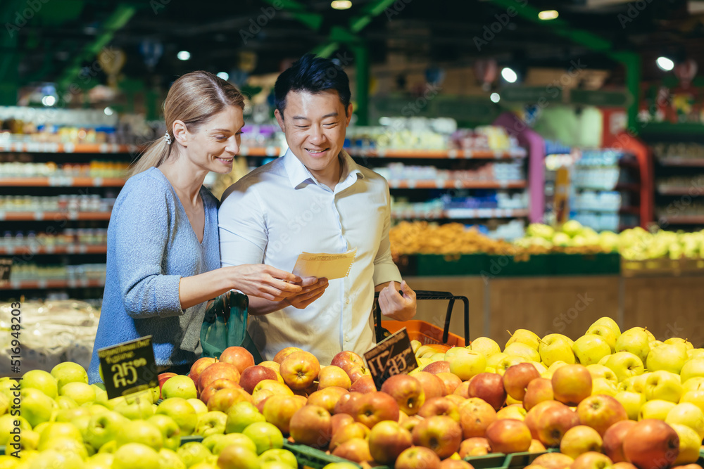 happy mixed race asian couple family man and woman choosing fruits or vegetables in grocery store supermarket. Joint daily shopping together. Buyers customer select product pick an apple in eco bag