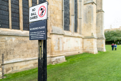Shallow focus of a No Ball Game and No Flying Drone sign seen outside a British cathedral. Visitors can be seen in the background, walking the perimeter.