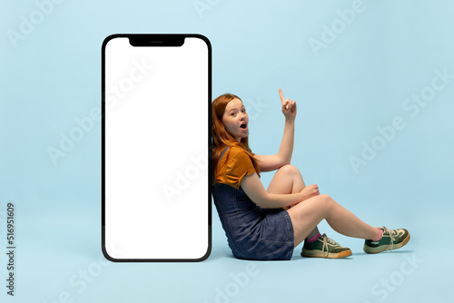 Young happy girl next to huge 3d model of smartphone with empty white screen isolated on blue background, Fashion, new app or website, copy space for your ad, mockup