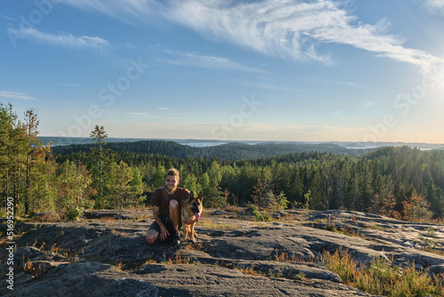 Dog is traveler. Ladoga Lake in distance and dense forest. Popular tourist destination. Hiidenvuori Mountain, Karelia. Man on hike with dog. Male owner next to German Shepherd sitting on top of cliff.