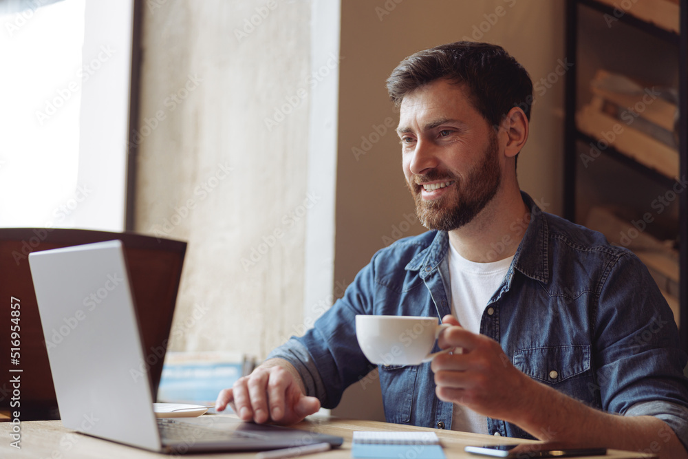 Portrait shot of cheerful male with beard holding cup of coffee, smiling in front of laptop screen. Portrait shot of cheerful male with beard holding cup of coffee, smiling in front of laptop screen.
