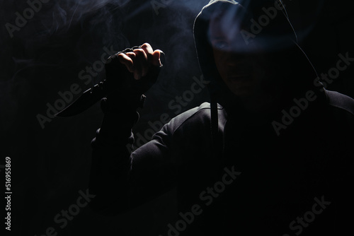 Silhouette of african american bandit in hood holding knife on black background with smoke