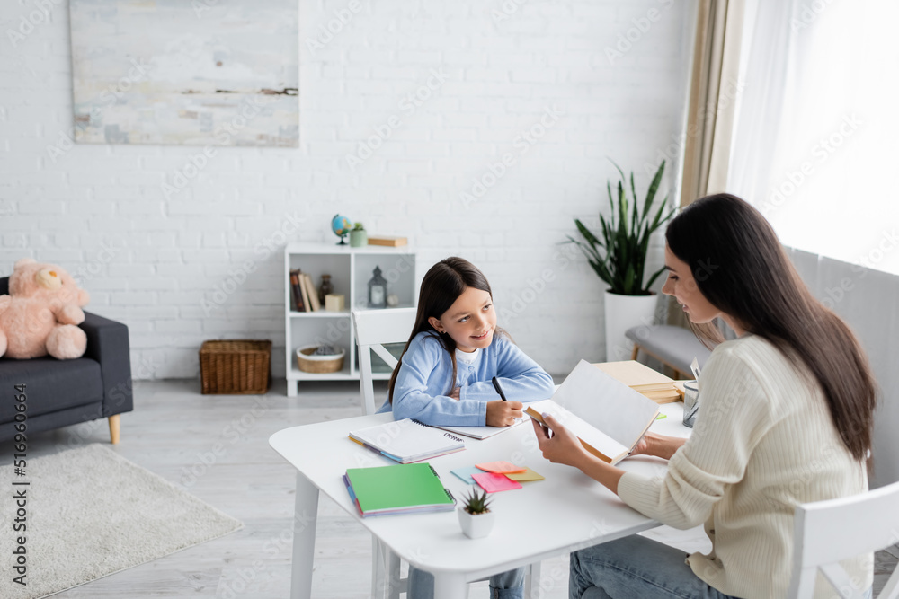 nanny reading book near smiling girl doing homework in modern living room. nanny reading book near smiling girl doing homework in modern living room.