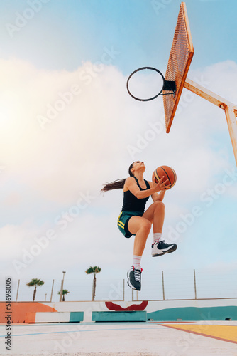 Low angle of woman playing basketball and jumping hoop against blue sky, copy space. Street game. Sport, practice and effort