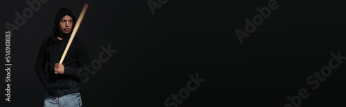 African american hooligan in hoodie holding baseball bat isolated on black, banner