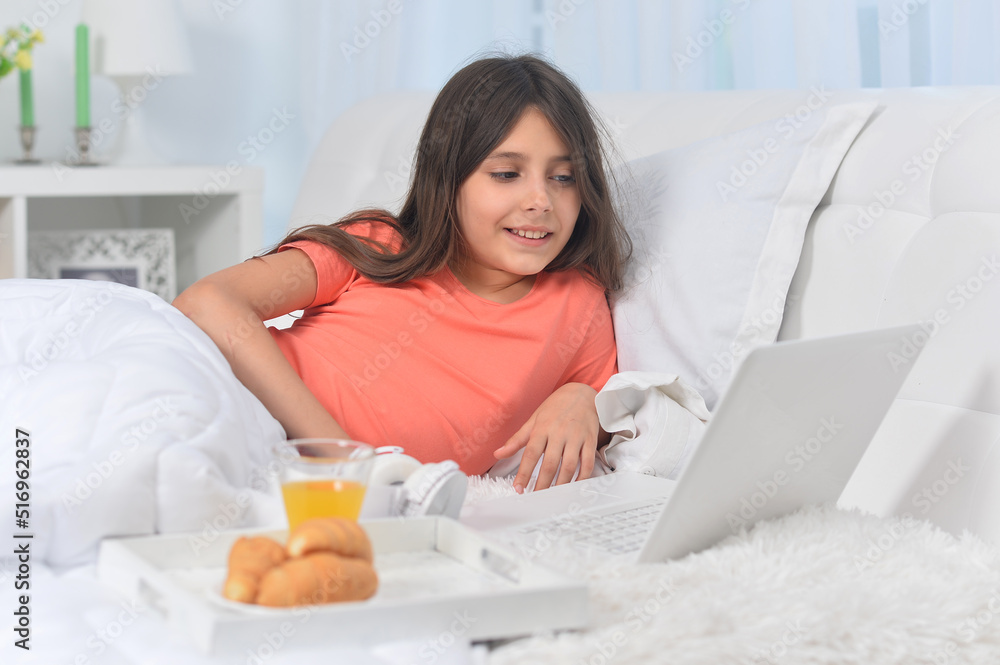 Beautiful girl  lying on soft beige bed with laptop and having breakfast