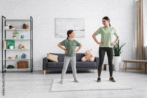 full length of positive nanny and child standing with hands on hips while exercising at home.