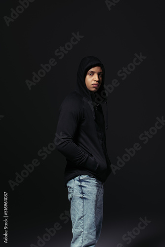 African american hooligan holding hand in pocket of hoodie isolated on black