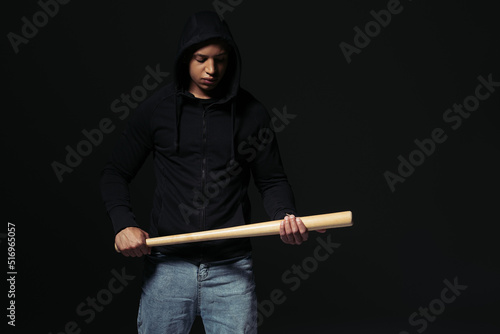 African american hooligan in hoodie looking at baseball bat isolated on black