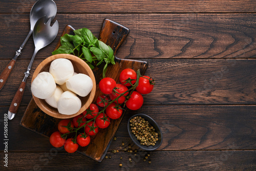 Caprese salad, ingredients for cooking. Cutting wooden board with traditional caprese preparation ingridients: mozzarella, tomatoes , basil, olive oil, cheese, spices on wooden rustic background.