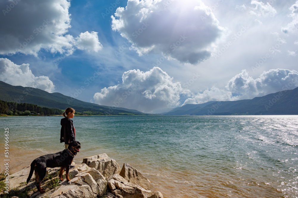 Girl stands near dog of Rottweiler breed on shore near lake against backdrop of mountain range covered with forest