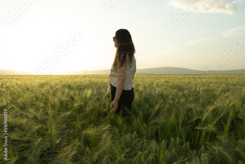 Woman farmer, young modern brunette woman farmer standing in the green wheat field and looking to sunset. Modern agriculture concept idea photo with copy space. Beautiful sunset in farmland.