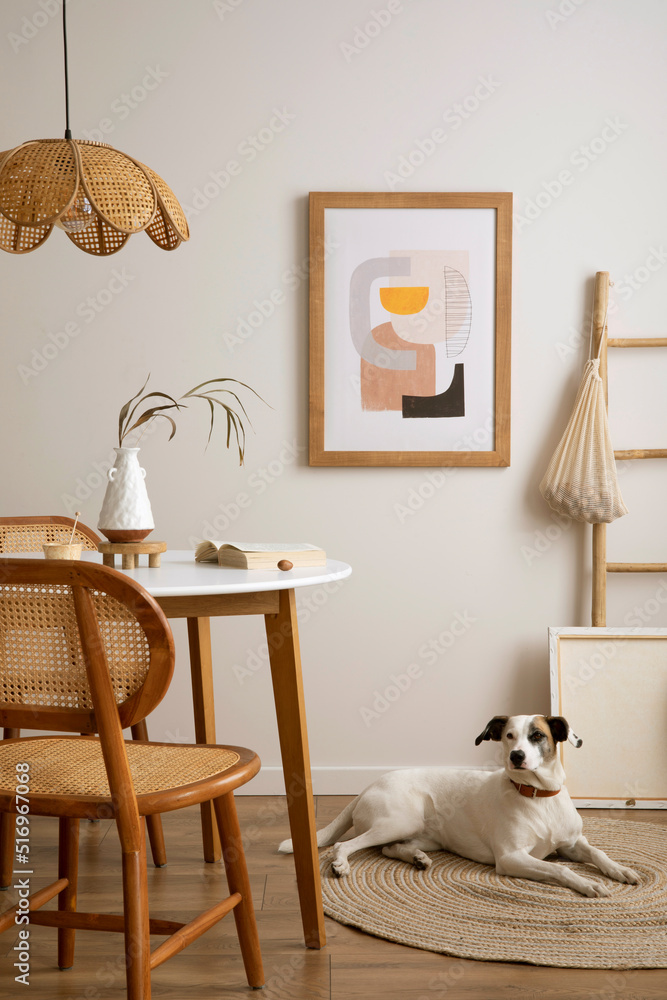 The stylish dining room with round table, rattan chair, dog on the carpet,  poster and kitchen accessories. Beige wall with mock up poster. Home decor.  Template. Stock Photo