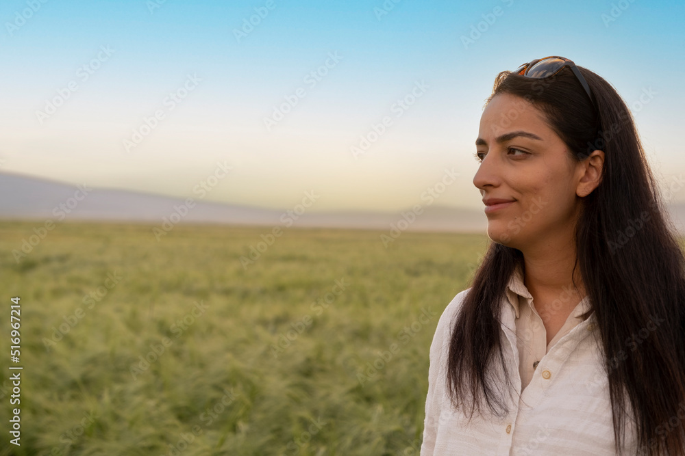 Agricultural wheat field, young farmer woman standing in front of agricultural wheat field in sunset. Beautiful agronomist farmer woman looking copy space area. Agriculture landscape concept idea.