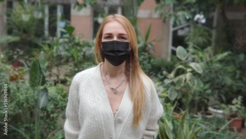 Portrait of young woman in medical mask standing against the background of plants in the greenhouse andBeautiful caucasian woman in black medical mask and white knitted sweater standing a in the photo