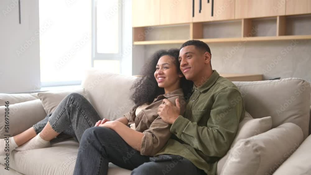 Side view of the multiracial couple chilling on the couch, watching movie in the studio apartment. Guy switching channel with TV remote Side view of the multiracial couple chilling on the couch, watching movie in the studio apartment. Guy switching channel with TV remote
