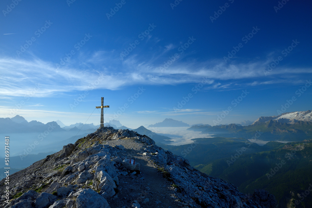 Das Gipfelkreuz des Sassongher in den Südtiroler Dolomiten am frühen morgen und im Hintergrund ist das Panorama, mit Gipfeln wie, Antelao, Pelmo, Civetta, und Marmolata zu sehen.