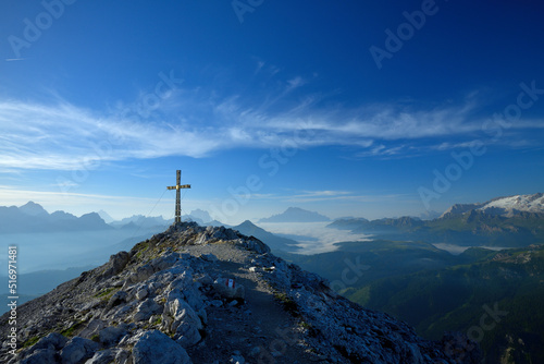 Das Gipfelkreuz des Sassongher in den Südtiroler Dolomiten am frühen morgen und im Hintergrund ist das Panorama, mit Gipfeln wie, Antelao, Pelmo, Civetta, und Marmolata zu sehen.