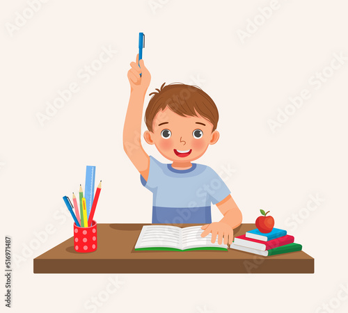Cute little boy student rising hand answering question sitting at her desk in the classroom