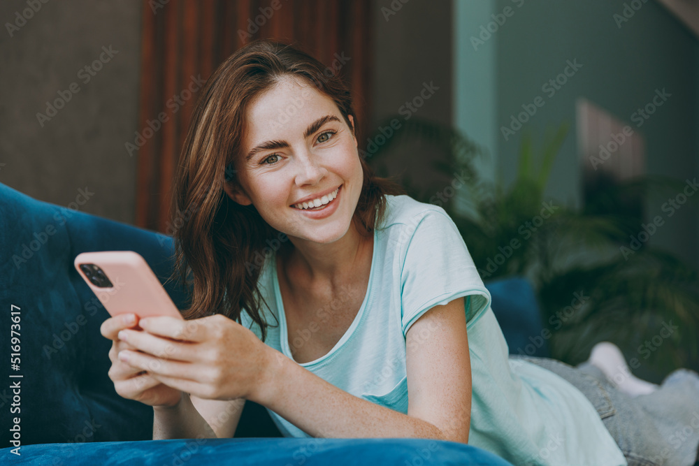 Young happy smiling cheerful woman she 20s in casual clothes mint t-shirt hold use mobile cell phone look camera lying on blue sofa indoor rest at home in own room apartment People lifestyle concept. Young happy smiling cheerful woman she 20s in casual clothes mint t-shirt hold use mobile cell phone look camera lying on blue sofa indoor rest at home in own room apartment People lifestyle concept.