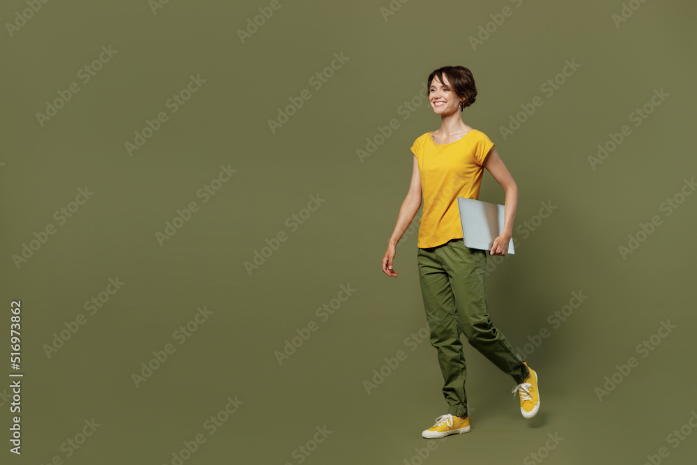 Full size side view young smiling happy woman she 20s wear yellow t-shirt hold closed laptop pc computer walk go isolated on plain olive green khaki background studio portrait People lifestyle concept Full size side view young smiling happy woman she 20s wear yellow t-shirt hold closed laptop pc computer walk go isolated on plain olive green khaki background studio portrait People lifestyle concept