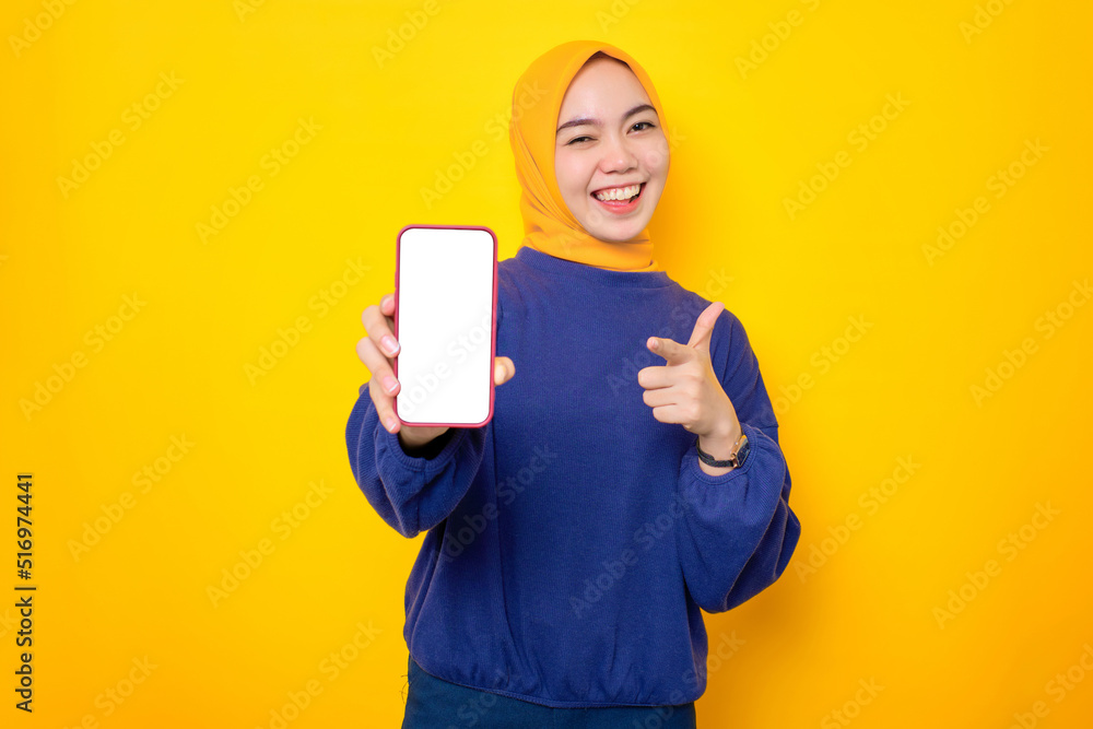 Excited young Asian Muslim woman dressed in casual sweater showing blank screen mobile phone, recommending mobile app isolated over yellow background Excited young Asian Muslim woman dressed in casual sweater showing blank screen mobile phone, recommending mobile app isolated over yellow background