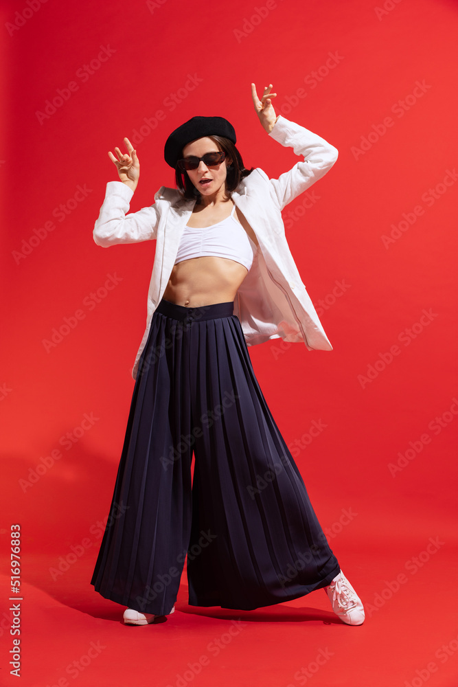 Stylish young woman in black beret and sunglasses posing, dancing isolated over red studio background. Parisian look Stylish young woman in black beret and sunglasses posing, dancing isolated over red studio background. Parisian look
